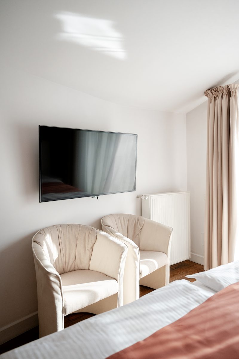 Dwa fotele pod telewizorem na ścianie, na pierwszym planie brzeg łóżka