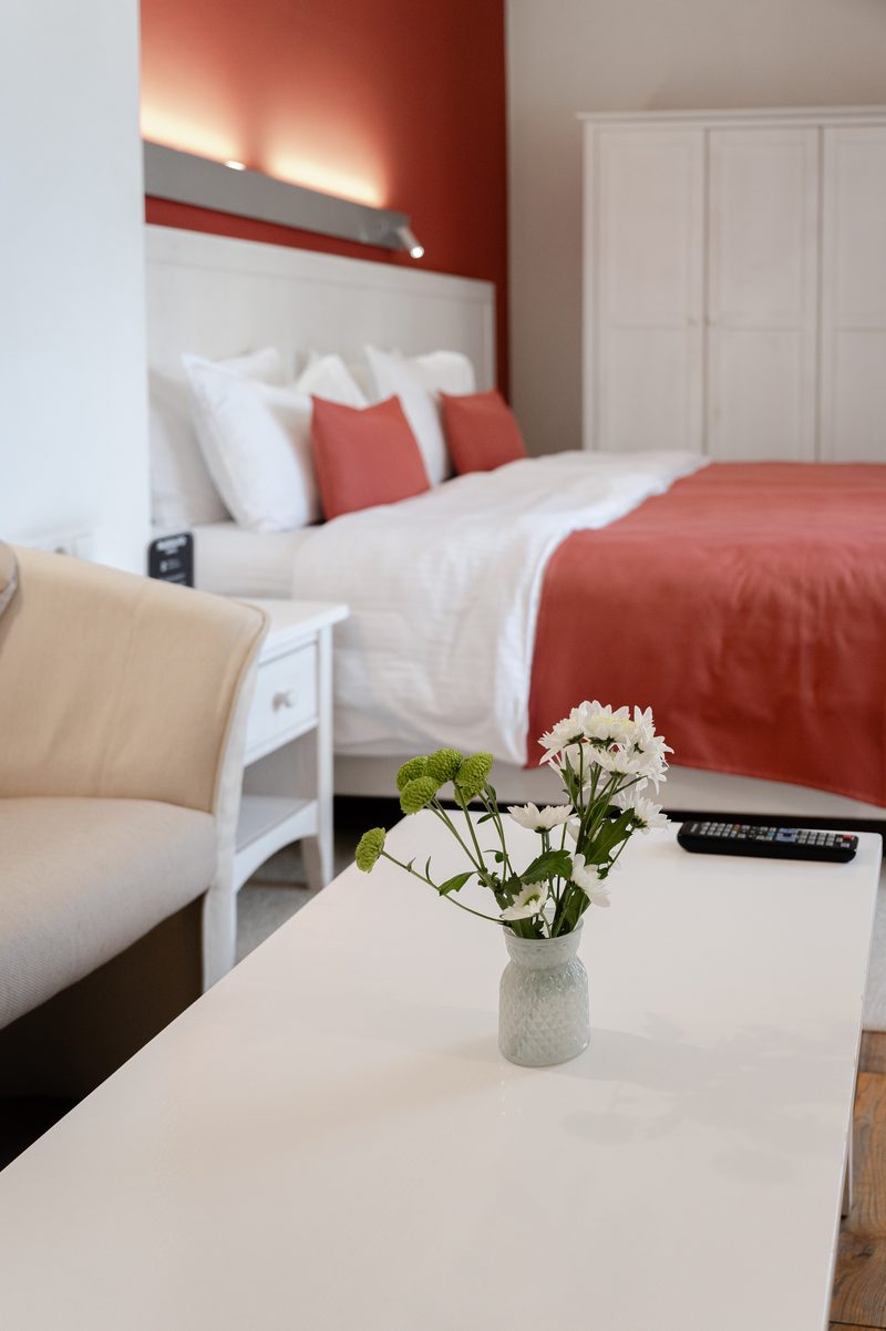 Das Bett mit einem roten Überwurf, davor Blumen auf dem Tisch